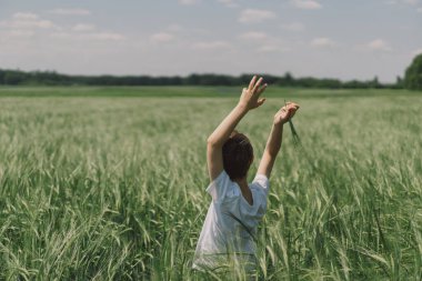 Beyaz tişörtlü ve yeşil arpa tarlasında oynayan gülümseyen bir çocuğun portresi. Mutlu çocuk yaz günü gülüyor ve oynuyor. Doğayı keşfeden çocuk. Arka plan