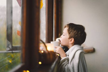 Battaniyeye sarılmış, sıcak çay içen ve sonbahar doğasına açık pencereden bakan sevimli bir çocuk. Sonbahar evi dekorasyonu. Samimi düşüş havası. Şükran Günü. Cadılar Bayramı.