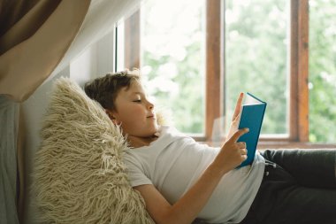 Pencerenin yanındaki pencere pervazında kitap okuyan sevimli çocuk. Güneşli bir günde evde dinlenen bir çocuk. Summer. Ev Okulu ve Eğitim Konsepti