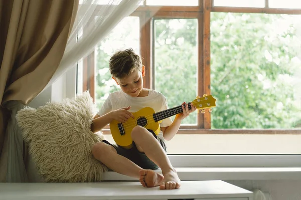 Tatlı çocuk pencerenin yanındaki pencere pervazında sarı Ukulele gitarı çalmayı öğreniyor. Rahat bir ev. Yaz tatili yaşam tarzı.