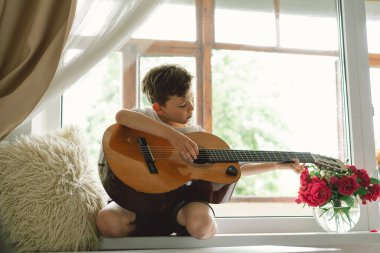 Tatlı çocuk pencerenin yanındaki pencere pervazında klasik gitar çalmayı öğreniyor. Rahat bir ev. Yaz tatili yaşam tarzı.