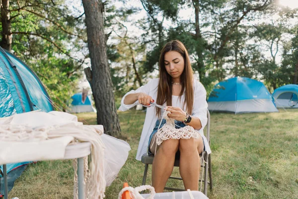 stock image Woman knits bag using macrame technique outdoors near tents. Outdoor hobbies. Macrame weaving.