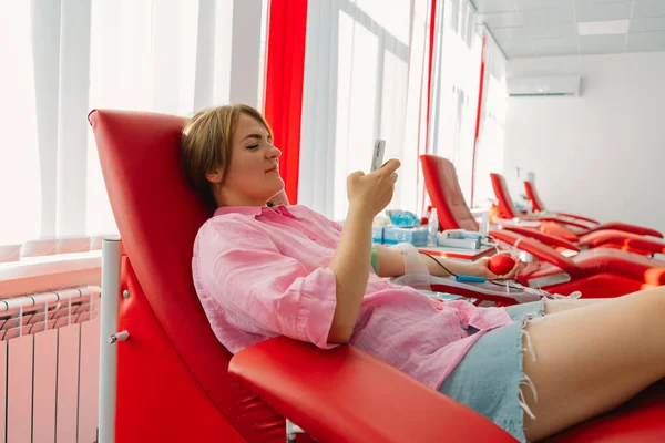 stock image Young woman with a phone making blood donation in hospital. A woman donates blood while holding a red heart. Blood donation.