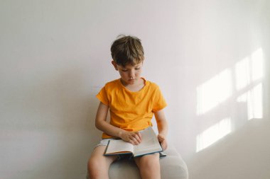 Turuncu tişört giyen sevimli bir çocuk yumuşak bir sedirin üzerinde oturmuş kitap okuyor. Uzayı kopyala Gerçek insanlar ve yaşam tarzı