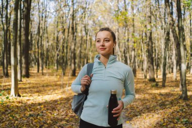 Kız demir şişeden su içiyor, plastikten değil. Güzel bir kız güneşli bir sonbahar ormanında dışarıda spor yapıyor. Vücut pozitifliği, kadınlar için spor, sağlıklı yaşam tarzı, kendini sevme ve sağIık.