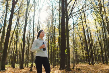 Kız demir şişeden su içiyor, plastikten değil. Güzel bir kız güneşli bir sonbahar ormanında dışarıda spor yapıyor. Vücut pozitifliği, kadınlar için spor, sağlıklı yaşam tarzı, kendini sevme ve sağIık.