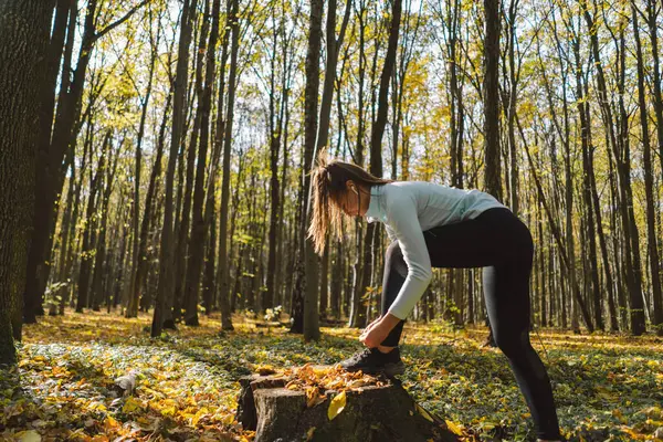The girl ties her shoelaces. Beautiful girl doing fitness in nature on a sunny autumn forest. Body positive, sports for women, harmony, healthy lifestyle, self-love and wellness.