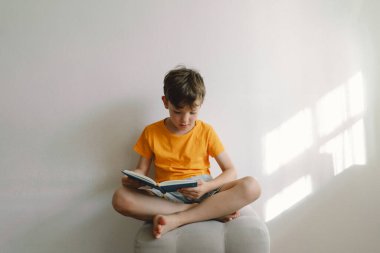 Turuncu tişört giyen sevimli bir çocuk yumuşak bir sedirin üzerinde oturmuş kitap okuyor. Uzayı kopyala Gerçek insanlar ve yaşam tarzı