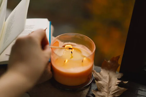 Hand with burning match lighting a candle on the windowsill with cozy autumn still life with sweater, candle and autumn decor. Autumn home decor. Cozy fall mood.