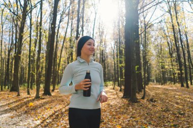 Kız demir şişeden su içiyor, plastikten değil. Güzel bir kız güneşli bir sonbahar ormanında dışarıda spor yapıyor. Vücut pozitifliği, kadınlar için spor, sağlıklı yaşam tarzı, kendini sevme ve sağIık.