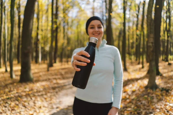 The girl drinks water from an iron bottle, not plastic. A beautiful girl is doing fitness outdoors in a sunny autumn forest. Body positive, sports for women, healthy lifestyle, self-love and wellness.