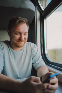 Mavi tişörtlü, sakallı ve bıyıklı bir adam trenle seyahat ederken akıllı telefon kullanıyor, trende oturuyor ve pencereden dışarı bakıyor..
