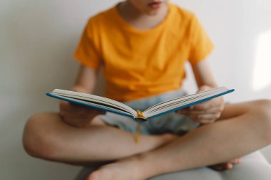 Turuncu tişört giyen sevimli bir çocuk yumuşak bir sedirin üzerinde oturmuş kitap okuyor. Uzayı kopyala Gerçek insanlar ve yaşam tarzı