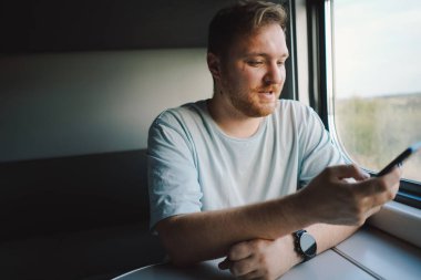 Mavi tişörtlü, sakallı ve bıyıklı bir adam trenle seyahat ederken akıllı telefon kullanıyor, trende oturuyor ve pencereden dışarı bakıyor..