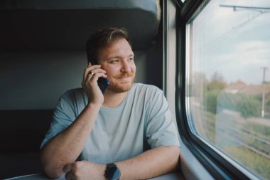 Mavi tişörtlü, sakallı ve bıyıklı bir adam trenle seyahat ederken akıllı telefon kullanıyor, trende oturuyor ve pencereden dışarı bakıyor..