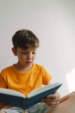 Turuncu tişört giyen sevimli bir çocuk yumuşak bir sedirin üzerinde oturmuş kitap okuyor. Uzayı kopyala Gerçek insanlar ve yaşam tarzı