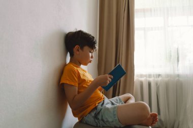 Turuncu tişört giyen sevimli bir çocuk yumuşak bir sedirin üzerinde oturmuş kitap okuyor. Uzayı kopyala Gerçek insanlar ve yaşam tarzı