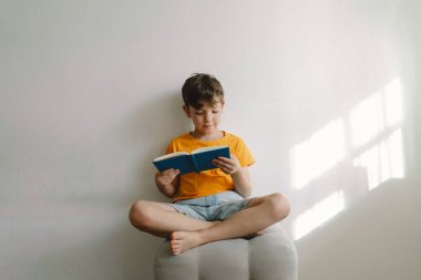 Turuncu tişört giyen sevimli bir çocuk yumuşak bir sedirin üzerinde oturmuş kitap okuyor. Uzayı kopyala Gerçek insanlar ve yaşam tarzı