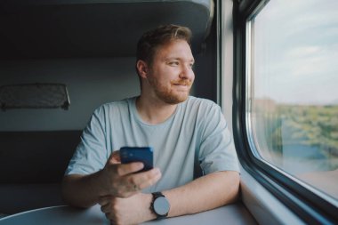 Mavi tişörtlü, sakallı ve bıyıklı bir adam trenle seyahat ederken akıllı telefon kullanıyor, trende oturuyor ve pencereden dışarı bakıyor..