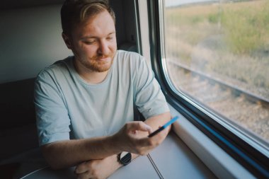 Mavi tişörtlü, sakallı ve bıyıklı bir adam trenle seyahat ederken akıllı telefon kullanıyor, trende oturuyor ve pencereden dışarı bakıyor..