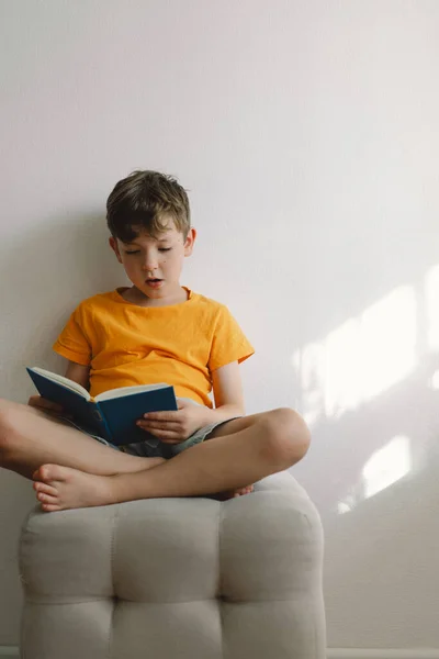 A cute boy wearing an orange T-shirt is sitting on a soft ottoman reading a book. Copy space. Real people and lifestyle