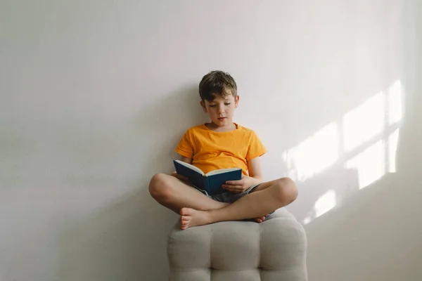 A cute boy wearing an orange T-shirt is sitting on a soft ottoman reading a book. Copy space. Real people and lifestyle