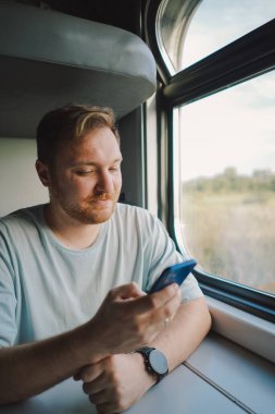 Mavi tişörtlü, sakallı ve bıyıklı bir adam trenle seyahat ederken akıllı telefon kullanıyor, trende oturuyor ve pencereden dışarı bakıyor..