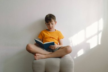 Turuncu tişört giyen sevimli bir çocuk yumuşak bir sedirin üzerinde oturmuş kitap okuyor. Uzayı kopyala Gerçek insanlar ve yaşam tarzı