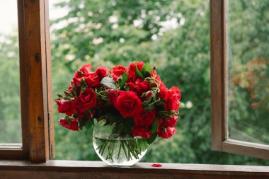 Vazoda bir buket kırmızı gül ve tahta pencere pervazında bir kitap. Eski bir kır evinin penceresinde, yazlık evde hala hayat var. Çiçekli ev dekorasyonu.