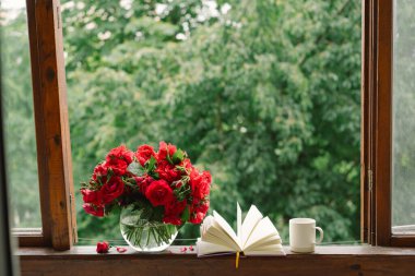 Vazoda bir buket kırmızı gül ve tahta pencere pervazında bir kitap. Eski bir kır evinin penceresinde, yazlık evde hala hayat var. Çiçekli ev dekorasyonu.