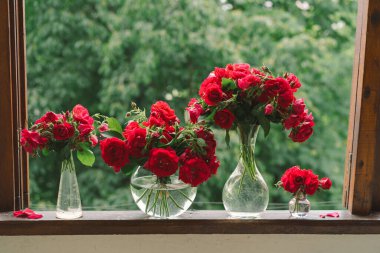 Vazoda bir buket kırmızı gül ve tahta pencere pervazında bir kitap. Eski bir kır evinin penceresinde, yazlık evde hala hayat var. Çiçekli ev dekorasyonu.
