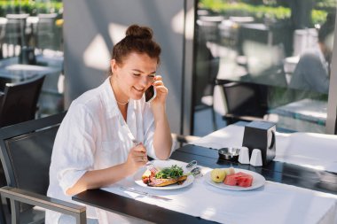 Bir kadın telefon kullanır ve bir kafede öğle veya dışarıda kahvaltı yapar. Restoran terasında sağlıklı yemek yiyen bir kadın..