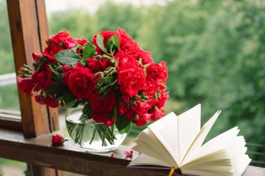 Vazoda bir buket kırmızı gül ve tahta pencere pervazında bir kitap. Eski bir kır evinin penceresinde, yazlık evde hala hayat var. Çiçekli ev dekorasyonu.