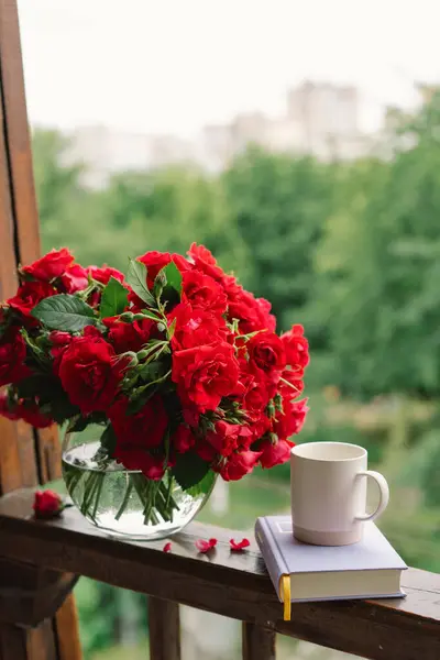 Vazoda bir buket kırmızı gül ve tahta pencere pervazında bir kitap. Eski bir kır evinin penceresinde, yazlık evde hala hayat var. Çiçekli ev dekorasyonu.