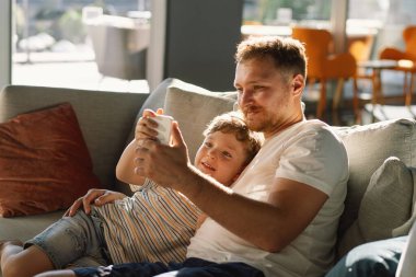 Baba ve oğul otel odasındaki kanepede rahatça oturup cep telefonuyla eğleniyorlar. Oğlan ve baba selfie telefonu yapıyor. Baba ve küçük çocuk kanepede dinleniyor ve akıllı telefondan komik videolar izliyorlar..