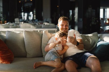 Baba ve oğul otel odasındaki kanepede rahatça oturup cep telefonuyla eğleniyorlar. Oğlan ve baba selfie telefonu yapıyor. Baba ve küçük çocuk kanepede dinleniyor ve akıllı telefondan komik videolar izliyorlar..