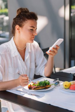 Bir kadın telefon kullanır ve bir kafede öğle veya dışarıda kahvaltı yapar. Restoran terasında sağlıklı yemek yiyen bir kadın..