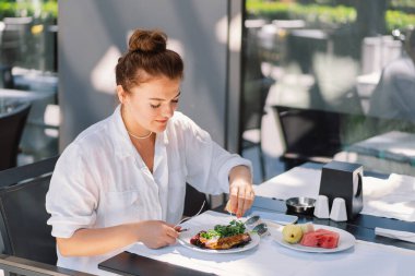 Beyaz gömlekli bir kadın bir kafede öğle yemeği ya da dışarıda kahvaltı yapar. Restoran terasında sağlıklı yemek yiyen bir kadın.