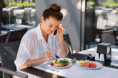 Bir kadın telefon kullanır ve bir kafede öğle veya dışarıda kahvaltı yapar. Restoran terasında sağlıklı yemek yiyen bir kadın..