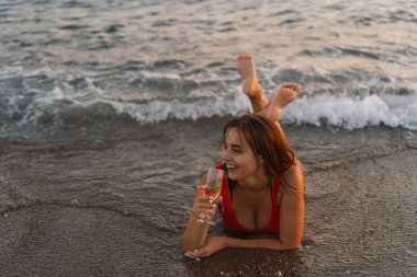 Canlı kırmızı mayo giymiş neşeli bir kadın elinde bir bardak beyaz şarapla deniz kenarında duruyor. Batan güneş, sahnenin üzerinde sıcak altın bir renk bırakıyor. Neşeli ifadesini vurguluyor..