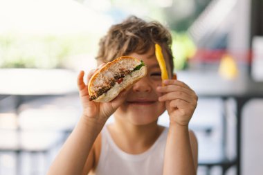 Masada sandviç ve patates kızartması yiyen küçük çocuk. Bir elinde bir sandviç, diğer elinde patates kızartmasıyla, yemeğine odaklanmış görünüyor. Bir çocuk dışarıda fast food yiyor..