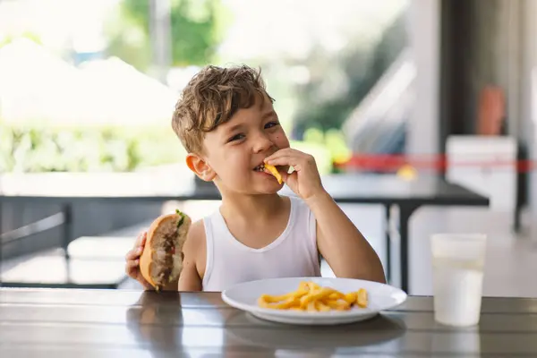 Pikkupoika Syö Voileipää Ranskalaisia Pöydässä Hän Näyttää Keskittynyt Hänen Ateria kuvapankin valokuva