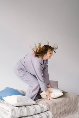 Neşeli bir kadın neşeli bir ifadeyle yastığı fırlatıyor. Etrafında bir yığın yastık ve yumuşak tekstil ile bej bir battaniyenin üzerinde çıplak ayakla duruyor. Rahat ve neşeli bir ortam öneriyor.