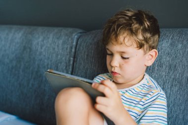 Genç bir çocuk gri bir kanepede oturuyor, tablet oynamaya derinden odaklanmış. Tişört giyiyor ve rahat görünüyor. Hareketlerine dalmış. İçerde sakin ve huzurlu bir öğleden sonra geçirdiğine işaret ediyor.