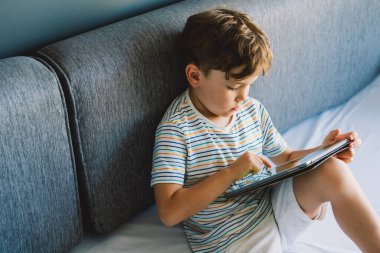 Genç bir çocuk gri bir kanepede oturuyor, tablet oynamaya derinden odaklanmış. Tişört giyiyor ve rahat görünüyor. Hareketlerine dalmış. İçerde sakin ve huzurlu bir öğleden sonra geçirdiğine işaret ediyor.