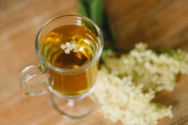 Elderflower çayı ile dolu berrak bir cam bardak kırsal bir ahşap masaya yerleştirilir. Taze mürver çiçekleri ve ahşap kesim tahtası yakınlarda, rahat ve doğal bir atmosfer yaratıyorlar..