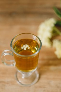 Elderflower çayı ile dolu berrak bir cam bardak kırsal bir ahşap masaya yerleştirilir. Taze mürver çiçekleri ve ahşap kesim tahtası yakınlarda, rahat ve doğal bir atmosfer yaratıyorlar..