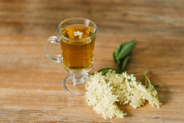 Elderflower çayı ile dolu berrak bir cam bardak kırsal bir ahşap masaya yerleştirilir. Taze mürver çiçekleri ve ahşap kesim tahtası yakınlarda, rahat ve doğal bir atmosfer yaratıyorlar..