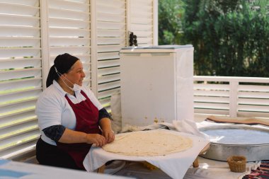 Bir kadın büyük, ince bir parça ekmek hazırlıyor. Bir açık hava pazarında çalışıyor, mutfak aletleri ve malzemeleriyle çevrili, yerel mutfak uygulamalarına bir göz atıyor..
