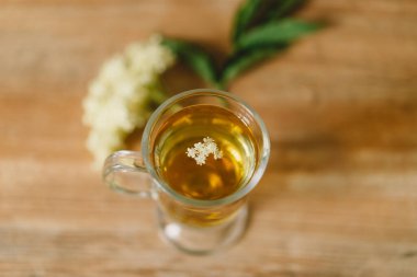 Elderflower çayı ile dolu berrak bir cam bardak kırsal bir ahşap masaya yerleştirilir. Taze mürver çiçekleri ve ahşap kesim tahtası yakınlarda, rahat ve doğal bir atmosfer yaratıyorlar..